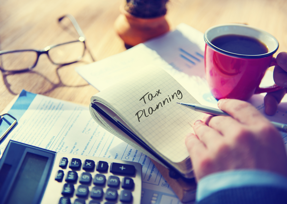 Businessman writing tax planning in notebook on desk with calculator, coffee, and paperwork