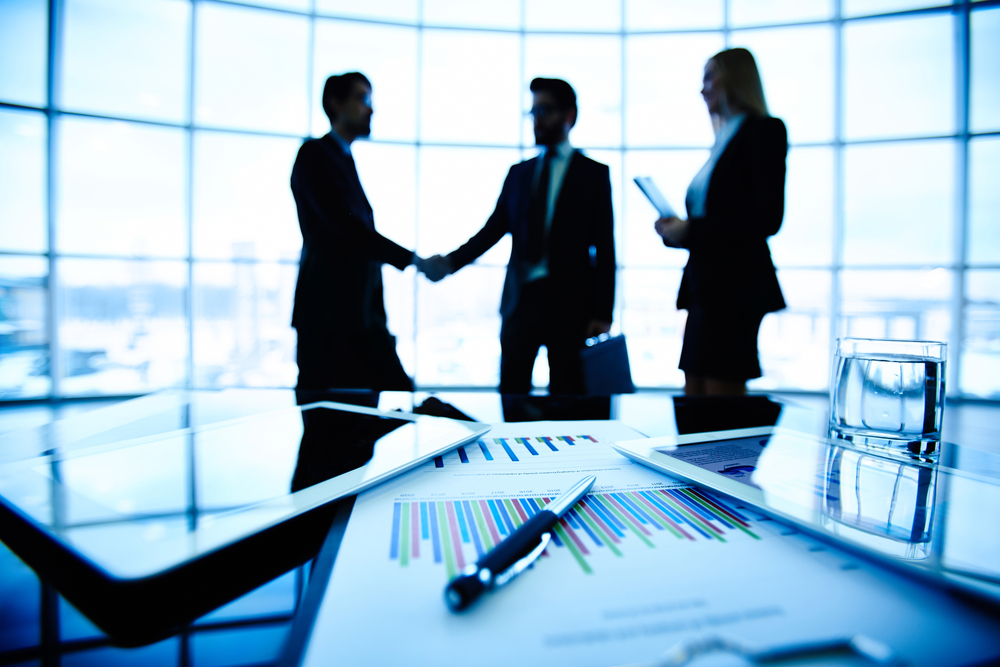 Silhouettes of 3 business professionals shaking hands with spreadsheets in foreground
